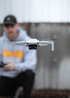 A close-up of a white drone in flight with blurred blades, centered in the foreground. In the background, a person wearing a black cap and gray hoodie appears softly out of focus, holding a controller.