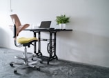 A workspace setup featuring a modern office chair with a yellow seat and pink backrest, positioned near a black desk made from an upcycled sewing machine base. On the desk, there is a closed laptop and a potted green plant, all against a plain white wall. The floor has a polished concrete finish.