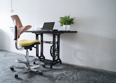 A workspace setup featuring a modern office chair with a yellow seat and pink backrest, positioned near a black desk made from an upcycled sewing machine base. On the desk, there is a closed laptop and a potted green plant, all against a plain white wall. The floor has a polished concrete finish.