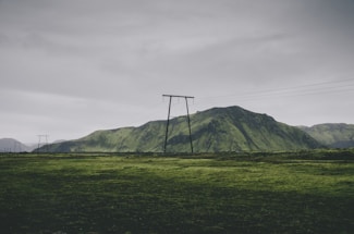 A landscape featuring a lush green field stretching towards a range of moss-covered hills. Power lines and poles cut across the scenery under an overcast sky, creating a contrast between nature and man-made structures.