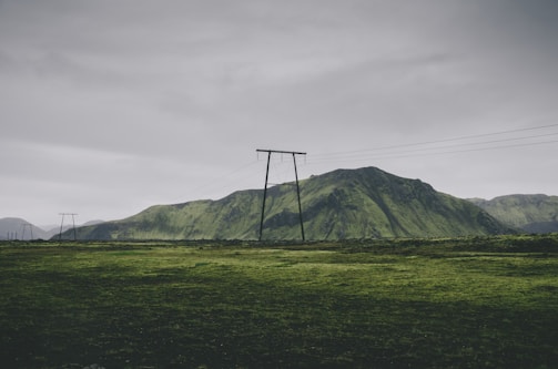 A landscape featuring a lush green field stretching towards a range of moss-covered hills. Power lines and poles cut across the scenery under an overcast sky, creating a contrast between nature and man-made structures.