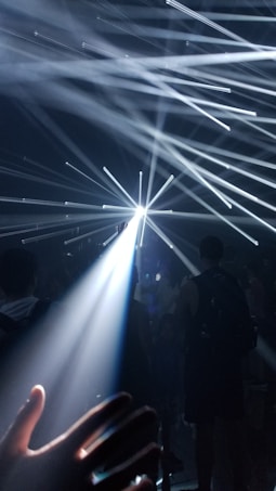 Beams of light radiate outward in a dark environment, creating a dramatic and dynamic effect. Silhouettes of people are visible, adding depth and context to the scene. A hand is in the foreground, reaching towards the light.