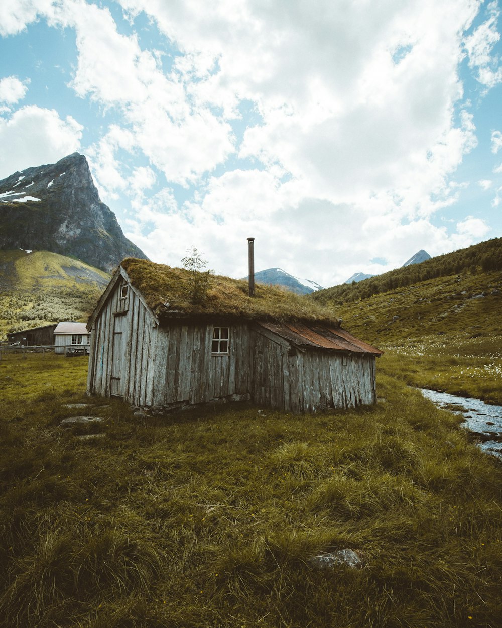 brown wooden house on green grass field near mountain under white clouds during daytime