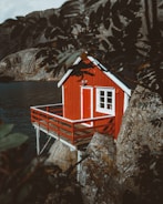 A small red cabin perched on wooden stilts is built against rocky terrain, overlooking a body of water. The scene is partially framed by leaves in the foreground, giving it a secluded and natural setting. The cabin features a white-framed door and windows, creating a striking contrast against the red exterior.