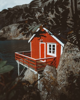 A small red cabin perched on wooden stilts is built against rocky terrain, overlooking a body of water. The scene is partially framed by leaves in the foreground, giving it a secluded and natural setting. The cabin features a white-framed door and windows, creating a striking contrast against the red exterior.