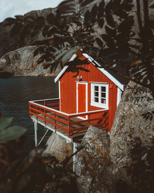 A small red cabin perched on wooden stilts is built against rocky terrain, overlooking a body of water. The scene is partially framed by leaves in the foreground, giving it a secluded and natural setting. The cabin features a white-framed door and windows, creating a striking contrast against the red exterior.