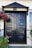 A polished black double door with a geometric panel design, adorned with a gold handle and knocker. The name 'Eriksen' is displayed on a plaque. A hanging basket filled with vibrant pink and red flowers shades the left side, providing a contrast to the serene and classic style of the door. White wood paneling surrounds the entrance.