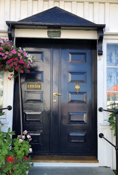 A polished black double door with a geometric panel design, adorned with a gold handle and knocker. The name 'Eriksen' is displayed on a plaque. A hanging basket filled with vibrant pink and red flowers shades the left side, providing a contrast to the serene and classic style of the door. White wood paneling surrounds the entrance.