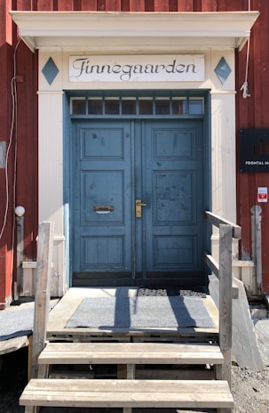 A double wooden door painted blue with a slightly worn appearance, flanked by a wooden staircase leading up to it. A sign above the door reads 'Finnegaarden' in elegant script. The building's facade is red with white trim, and a black sign is visible to the right.