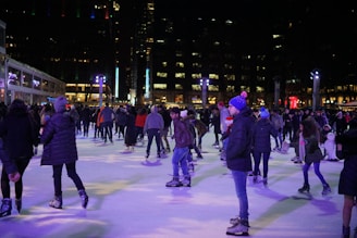 People are enjoying a nighttime skating session on a well-lit ice rink. The scene is bustling with activity as skaters of various ages move around the rink. Some people are standing, chatting, while others glide gracefully across the ice. Warm clothing, including hats and jackets, is common among the skaters. In the background, tall buildings with numerous lit windows intersperse with decorative lights creating a festive atmosphere.