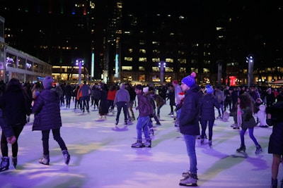 People are enjoying a nighttime skating session on a well-lit ice rink. The scene is bustling with activity as skaters of various ages move around the rink. Some people are standing, chatting, while others glide gracefully across the ice. Warm clothing, including hats and jackets, is common among the skaters. In the background, tall buildings with numerous lit windows intersperse with decorative lights creating a festive atmosphere.