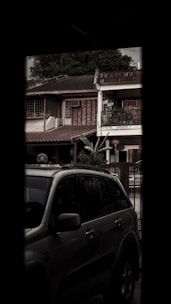 A partial view of a residential house with a tile roof and a balcony, partially obscured by the presence of a parked SUV in the foreground. There are trees in the background and various architectural details on the house that suggest a vintage aesthetic.