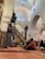 A group of children sit attentively on a carpet in a mosque, listening to a man standing before them. The architecture features high arched ceilings and ornate designs. The children are wearing colorful clothing, some with headscarves or masks.