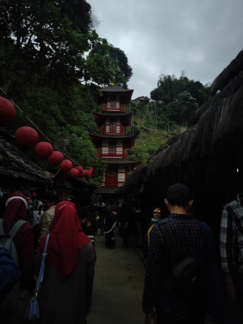 A tall pagoda structure with multiple tiers and red and white accents is surrounded by lush green vegetation. A pathway crowded with people leads to the pagoda, and red lanterns are strung across the area, creating a festive atmosphere. The sky is overcast, adding a moody ambiance.