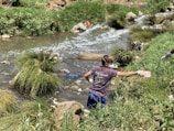 A person wearing gloves and a mask is cleaning up a littered stream surrounded by grass and rocks. The waterway is cluttered with various debris, indicating pollution.