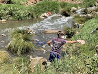 A person wearing gloves and a mask is cleaning up a littered stream surrounded by grass and rocks. The waterway is cluttered with various debris, indicating pollution.