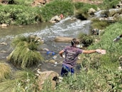 A person wearing gloves and a mask is cleaning up a littered stream surrounded by grass and rocks. The waterway is cluttered with various debris, indicating pollution.