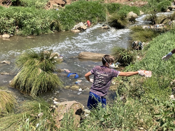 A person wearing gloves and a mask is cleaning up a littered stream surrounded by grass and rocks. The waterway is cluttered with various debris, indicating pollution.