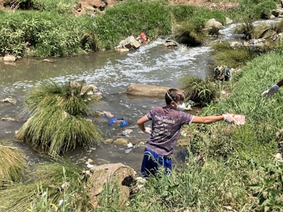 A person wearing gloves and a mask is cleaning up a littered stream surrounded by grass and rocks. The waterway is cluttered with various debris, indicating pollution.