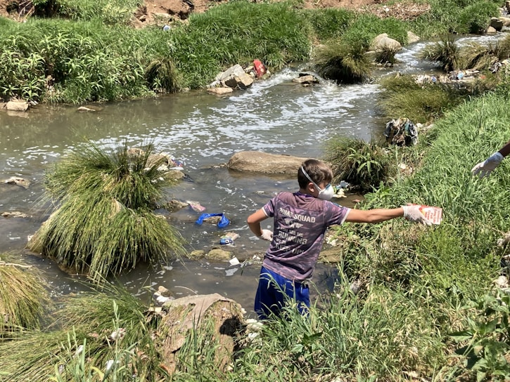 A person wearing gloves and a mask is cleaning up a littered stream surrounded by grass and rocks. The waterway is cluttered with various debris, indicating pollution.