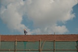 A person wearing a hat works on a tiled roof under a blue sky with large white clouds. The scene portrays typical rural or suburban architecture with a focus on the solitary worker who seems engaged in repairing or inspecting the roof tiles. Below the tiles, a semi-transparent green netting is visible, possibly for safety or construction purposes.