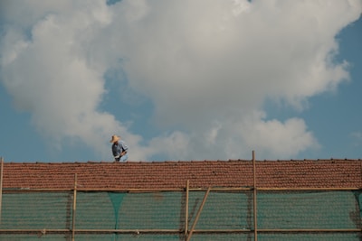 A person wearing a hat works on a tiled roof under a blue sky with large white clouds. The scene portrays typical rural or suburban architecture with a focus on the solitary worker who seems engaged in repairing or inspecting the roof tiles. Below the tiles, a semi-transparent green netting is visible, possibly for safety or construction purposes.