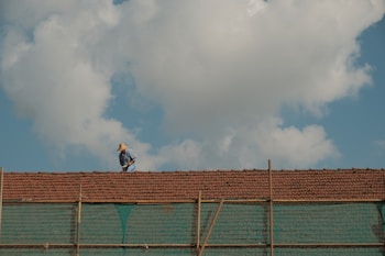A person wearing a hat works on a tiled roof under a blue sky with large white clouds. The scene portrays typical rural or suburban architecture with a focus on the solitary worker who seems engaged in repairing or inspecting the roof tiles. Below the tiles, a semi-transparent green netting is visible, possibly for safety or construction purposes.