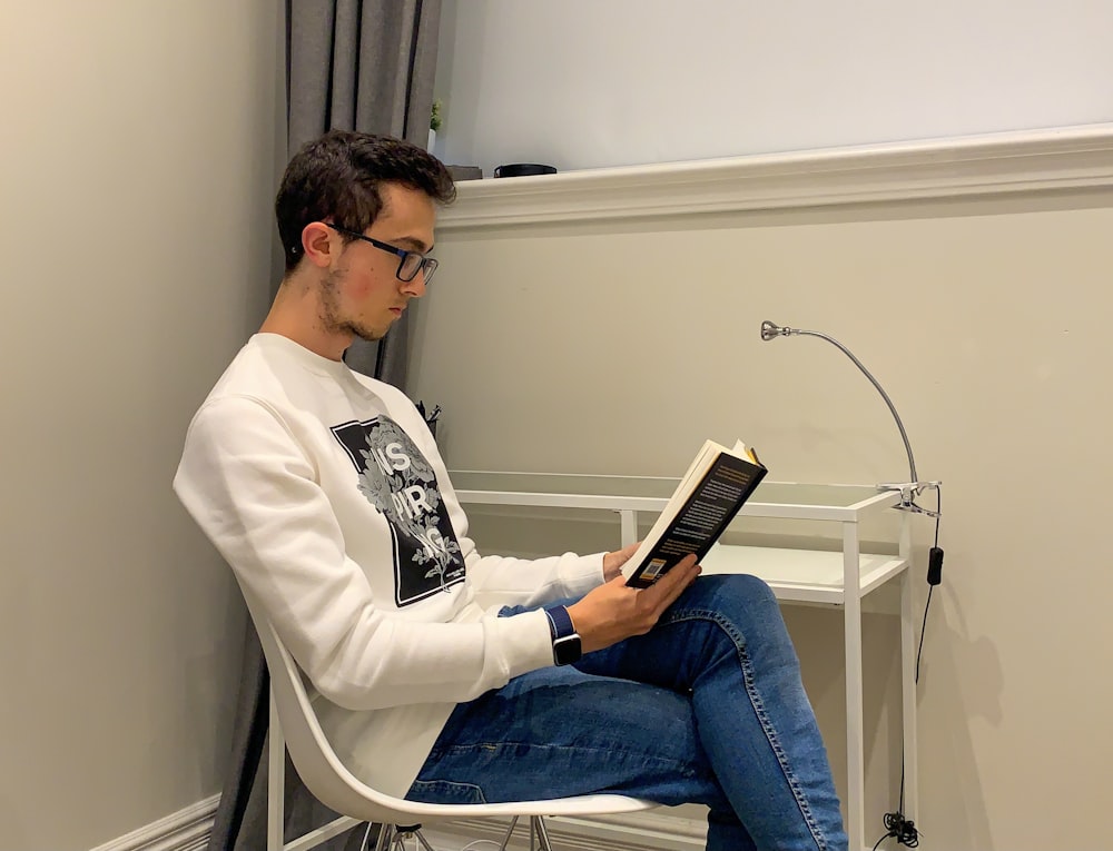 man in white dress shirt and blue denim jeans sitting on white chair reading book