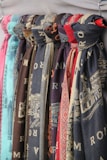 Multiple scarves are hanging in a row, displaying various patterns and colors. Each scarf appears to have printed designs, some with textual elements and others with intricate patterns. The colors of the scarves range from shades of blue and red to brown and teal.
