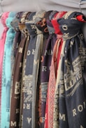 Multiple scarves are hanging in a row, displaying various patterns and colors. Each scarf appears to have printed designs, some with textual elements and others with intricate patterns. The colors of the scarves range from shades of blue and red to brown and teal.