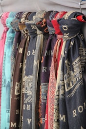 Multiple scarves are hanging in a row, displaying various patterns and colors. Each scarf appears to have printed designs, some with textual elements and others with intricate patterns. The colors of the scarves range from shades of blue and red to brown and teal.