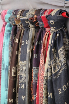 Multiple scarves are hanging in a row, displaying various patterns and colors. Each scarf appears to have printed designs, some with textual elements and others with intricate patterns. The colors of the scarves range from shades of blue and red to brown and teal.
