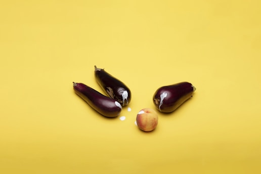 Three eggplants and a peach are placed on a bright yellow background. The eggplants form a triangular arrangement around the peach, and there is a small amount of white liquid splattered around them.