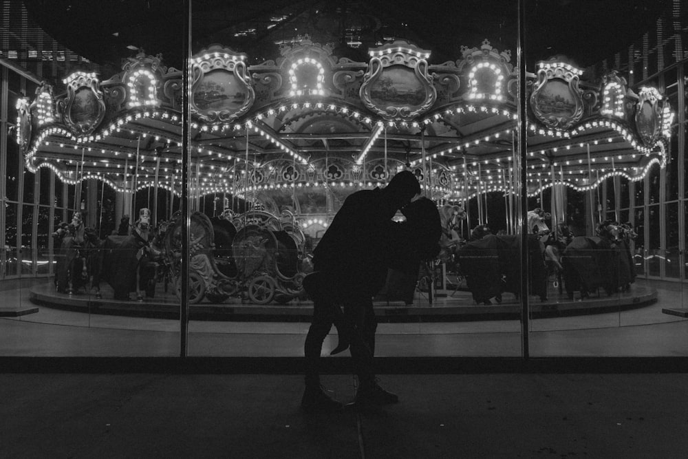 grayscale photo of a person in black jacket and pants standing in front of a carousel
