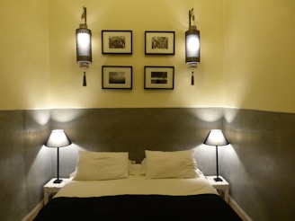 A neatly arranged bedroom with a double bed, adorned with white pillows and a dark bedspread. The bed is flanked by two small side tables, each with a modern table lamp emitting soft light. Above the bed, four framed black and white photographs are symmetrically placed. On each side, ornate wall-mounted lights provide a warm glow, adding a touch of elegance to the room. The walls have a two-tone design with a lighter top and a darker bottom.