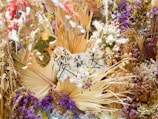A vibrant and intricate arrangement of dried flowers and foliage in various shades of purple, brown, and cream. There is a central focus on fans of dried palm leaves, surrounded by clusters of small flowers and leaves. Japanese characters are written on a piece of white material in the middle, adding a cultural element to the display.