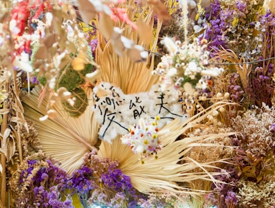 A vibrant and intricate arrangement of dried flowers and foliage in various shades of purple, brown, and cream. There is a central focus on fans of dried palm leaves, surrounded by clusters of small flowers and leaves. Japanese characters are written on a piece of white material in the middle, adding a cultural element to the display.