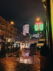 A nighttime urban street scene featuring wet pavements and reflections from streetlights and passing cars. Bright digital signage displays currency exchange rates in green and red. The street is lined with older buildings, and a modern skyscraper with illuminated patterns is visible in the background. Pedestrians and vehicles are moving along the street, with some people carrying umbrellas.