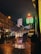A nighttime urban street scene featuring wet pavements and reflections from streetlights and passing cars. Bright digital signage displays currency exchange rates in green and red. The street is lined with older buildings, and a modern skyscraper with illuminated patterns is visible in the background. Pedestrians and vehicles are moving along the street, with some people carrying umbrellas.
