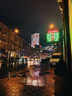 A nighttime urban street scene featuring wet pavements and reflections from streetlights and passing cars. Bright digital signage displays currency exchange rates in green and red. The street is lined with older buildings, and a modern skyscraper with illuminated patterns is visible in the background. Pedestrians and vehicles are moving along the street, with some people carrying umbrellas.