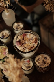 A collection of artisan candles with dried flowers and herbs embedded in the wax is arranged on a wooden surface. The candles vary in size, with some in glass containers. The dried flowers include lavender and rose petals, adding a rustic and handmade aesthetic. Soft lighting enhances the warm and cozy atmosphere.