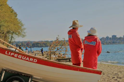 Two people wearing red lifeguard jackets and straw hats stand on a beach, facing the water. A lifeguard boat is visible in the foreground. In the background, there are trees, several people near the water's edge, and buildings on the distant shoreline.