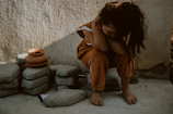 A young child sits on concrete blocks with their head resting on their arms in a corner. The child is wearing orange-brown clothing with white lace detailing, and their long hair is loose. Next to the child are stacked clay pots and more concrete blocks. The lighting casts soft shadows on the textured wall and ground.