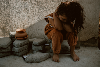 A young child sits on concrete blocks with their head resting on their arms in a corner. The child is wearing orange-brown clothing with white lace detailing, and their long hair is loose. Next to the child are stacked clay pots and more concrete blocks. The lighting casts soft shadows on the textured wall and ground.