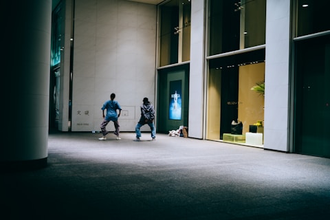 Two people are engaging in a dance or exercise routine in front of large glass windows. One person is wearing a blue shirt and patterned pants, while the other wears a jacket and darker pants. The setting appears to be a commercial space, possibly a mall or shopping area, with advertisements visible through the windows.