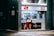 A small storefront that appears to be a tobacco shop with glass display windows showcasing various cigarette packs and tobacco products. The exterior is brightly lit, featuring posters with promotional images and text in Japanese. The interior is visible with shelves stocked with products, and a person inside attending to the store.