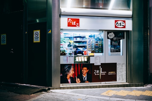 A small storefront that appears to be a tobacco shop with glass display windows showcasing various cigarette packs and tobacco products. The exterior is brightly lit, featuring posters with promotional images and text in Japanese. The interior is visible with shelves stocked with products, and a person inside attending to the store.
