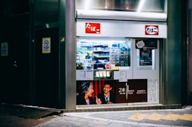 A small storefront that appears to be a tobacco shop with glass display windows showcasing various cigarette packs and tobacco products. The exterior is brightly lit, featuring posters with promotional images and text in Japanese. The interior is visible with shelves stocked with products, and a person inside attending to the store.