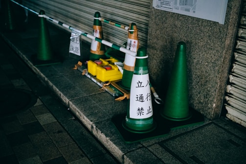 Several green traffic cones are placed along a sidewalk next to a metal barrier in an urban setting. The cones have Japanese writing on them, and there is a yellow weight placed on the ground, likely used to secure the barrier. The area appears to be cordoned off, indicating a no-entry zone.