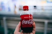 A hand is holding a small plastic bottle of Coca-Cola with a red label prominently displaying the brand name. The background is blurred, focusing attention on the bottle.